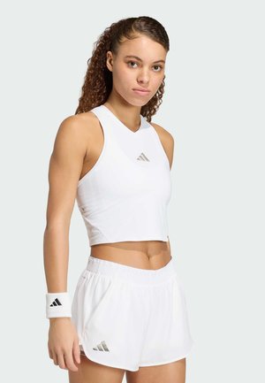 Młoda kobieta ubrana w biały bezrękawnik typu crop top, białe sportowe szorty oraz opaskę na nadgarstek z logo Adidas na jasnym tle.