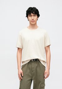 Béžové bavlněné tričko s kulatým výstřihem a krátkými rukávy, nošené s olivovými cargo kalhotami a černým opaskem se stříbrnými kovovými prvky.