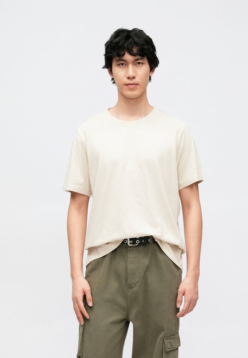 Béžové bavlněné tričko s kulatým výstřihem a krátkými rukávy, nošené s olivovými cargo kalhotami a černým opaskem se stříbrnými kovovými prvky.