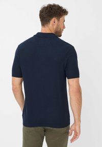 Navy Polo-Shirt mit kurzen Ärmeln und strukturiertem Finish. Hat einen klassischen Kragen und einen geraden Saum. Getragen mit khakifarbenen Hosen.