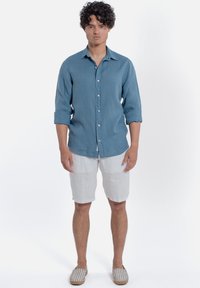Camicia azzurra chiaro, con bottoni, a maniche lunghe in misto lino, abbinata a pantaloni corti bianchi in lino. Il modello indossa espadrillas a righe. Sfondo bianco.