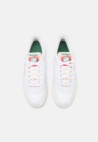 Um par de ténis brancos da Reebok com palmilhas verdes, área do dedo perfurada e patches de logo vermelhos nas línguas, vistos de cima sobre um fundo simples.