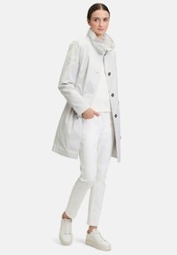 Lichtgrijze waterdichte trenchcoat met een hoge kraag, knoopsluiting, zijzakken en een relaxed fit, gecombineerd met witte broek en sneakers.