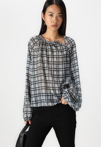 Blusa leggera e trasparente con un motivo a quadri nero e grigio, colletto arricciato e maniche lunghe; indossata con pantaloni neri e accessorata con una borsa.
