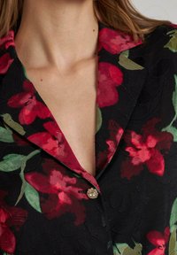 Blazer floral en tela negra, con patrones de flores rojas y verdes. Detalle notable de botones dorados con superficie texturizada.