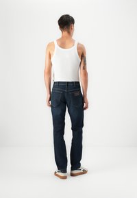 Jeans de mezclilla azul oscuro con un corte recto, bolsillos traseros y un parche de cuero. El modelo lleva una camiseta sin mangas blanca y zapatillas.