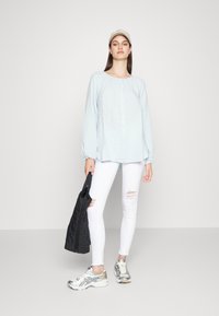 Camisa azul claro de botão, com mangas longas, jeans brancos rasgados, bolsa preta e ténis prateados, contra um fundo simples.