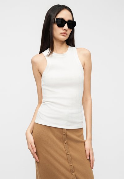 Λευκό ριμπ αμάνικο τοπ και φούστα σε ταμπέ απόχρωση με λεπτομέρειες κουμπιών. Το μοντέλο φοράει μαύρα τετράγωνα γυαλιά ηλίου, με μακριά και ίσια μαλλιά.