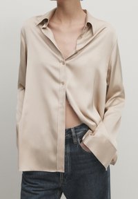 Personne portant un chemisier beige en satin surdimensionné partiellement déboutonné, avec un jean bleu foncé et une main dans la poche, sur un fond uni.