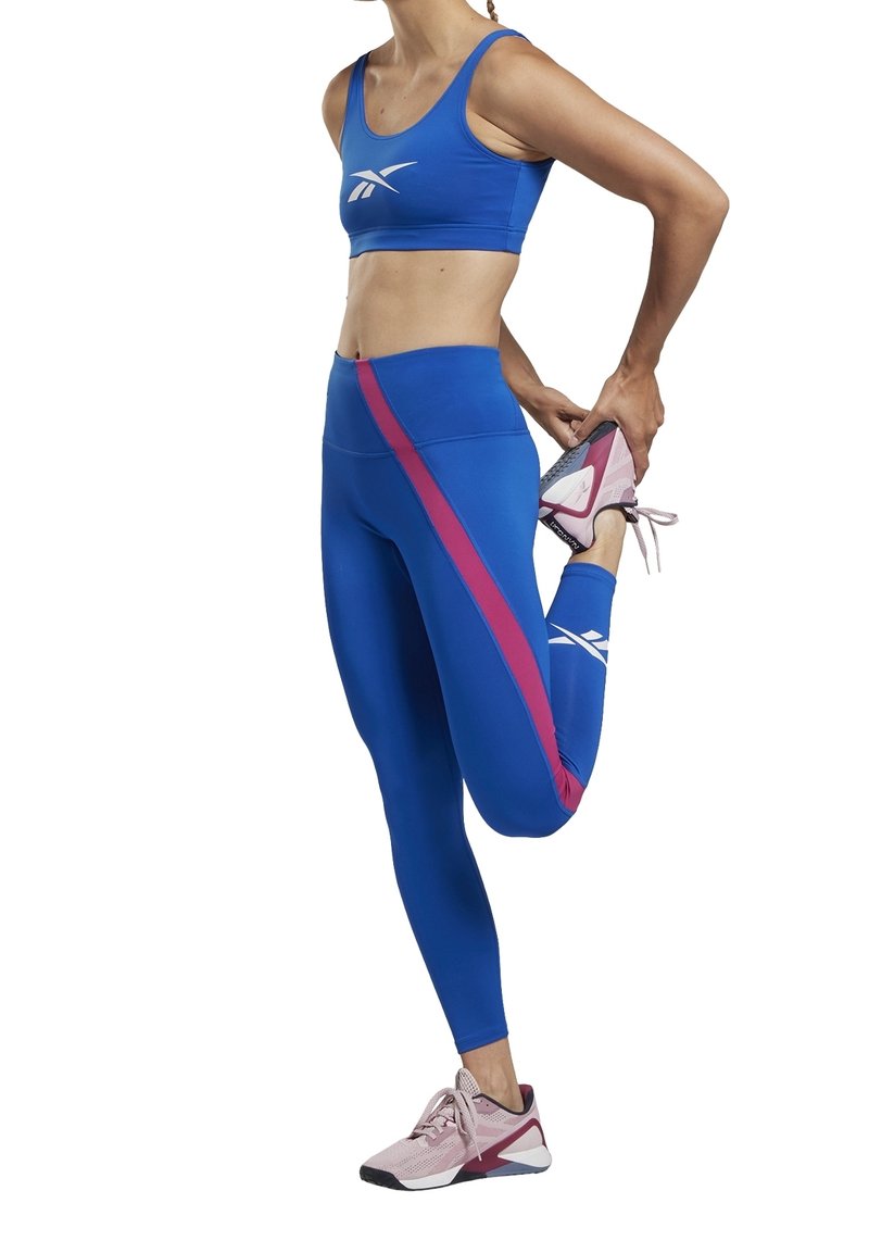 Modrý sportovní crop top a legíny s vysokým pasem s růžovým bočním pruhem; model provádí strečink; růžové sportovní boty s bílými akcenty.