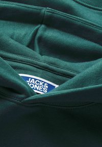 Primo piano del tessuto verde scuro di una felpa con cappuccio che mostra cuciture ricamate e un'etichetta del marchio "Jack & Jones" parzialmente visibile all'interno del colletto.
