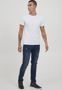 T-shirt bianca in cotone, jeans blu con effetto usurato e sneaker nere. Il modello è in piedi di lato, mostrando uno stile semplice e casual.