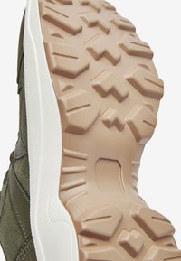 Sole od plátěných tenisek v hnědé gumě, s geometrickými vzory na podrážce pro lepší trakci. Boční pohled zobrazuje zelený textilní svršek a bílou mezipodešev.