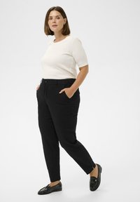 Sieviete stāv ar rokām melno bikses kabatās, tērpta baltā īsajās piedurknēs augšpusē un melnās loafers, pret vienkāršu baltu fonu.