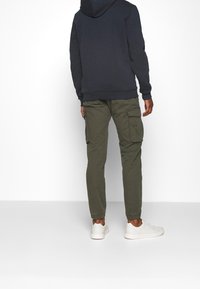 Felpa con cappuccio blu scuro, pantaloni cargo verdi oliva con tasche laterali e sneakers bianche. Il materiale sembra morbido e informale.
