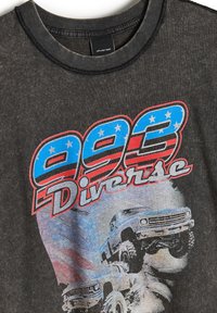 Czarny t-shirt z grafiką z czerwonym i niebieskim napisem "993 Diverse", z ilustracjami ciężarówek i wyblakłym wzorem. Okrągły dekolt i krótkie rękawy.