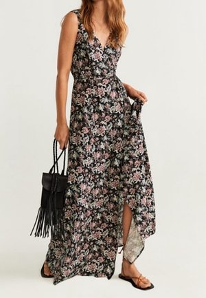 Kvinde i sort blomstermaxikjole uden ærmer med sidelomme, der holder en sort frynset håndtaske og har brune sandaler med stropper på.