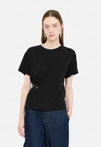 Juodas medvilninis T-shirt, turintis mazgo dizainą, pritvirtintą sidabriniu dirželiu, trumpomis rankovėmis ir laisvu siluetu. Dėvimas su tamsiomis džinsinėmis kelnėmis.
