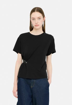 T-shirt in cotone nero con un design annodato fissato con una fibbia argentata, maniche corte e una vestibilità rilassata. Indossato con pantaloni in denim scuri.
