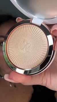 Compacte highlighterpoeder met zonnestraalpatroon en gegraveerde initialen, dicht bij het gezicht van een persoon met lange, verzorgde nagels gehouden.