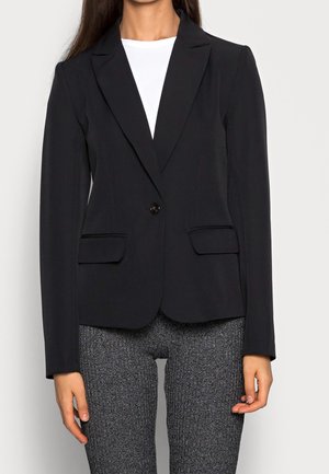 Vrouw draagt een nauwsluitend zwart colbert over een witte top en grijze, gestructureerde broek, staand tegen een effen lichte achtergrond.