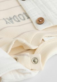 Camisa color crema con rayas marrón claro, que presenta una tapeta de botones, detalles de textura en la tela y broches de metal en la parte inferior.