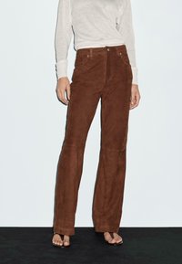 Brązowe zamszowe spodnie w szerokim kroju mają wysoki stan, zamek z przodu oraz metalowe akcenty w postaci guzików. W zestawie z jasnoszarym, długim rękawem.