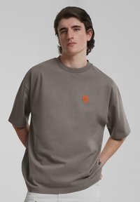 Camiseta gris de manga corta hecha de algodón con un corte holgado, que presenta un logo bordado naranja en el pecho. Textura suave, diseño de cuello redondo.