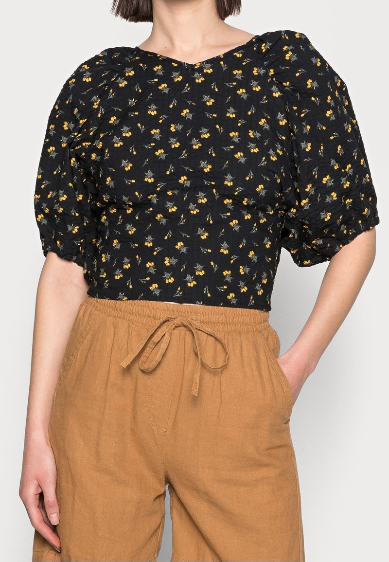 Blusa nera floreale con maniche a sbuffo, caratterizzata da motivi gialli e verdi. Abbinata a pantaloni corti elasticizzati di colore marrone chiaro. Tessuto morbido.