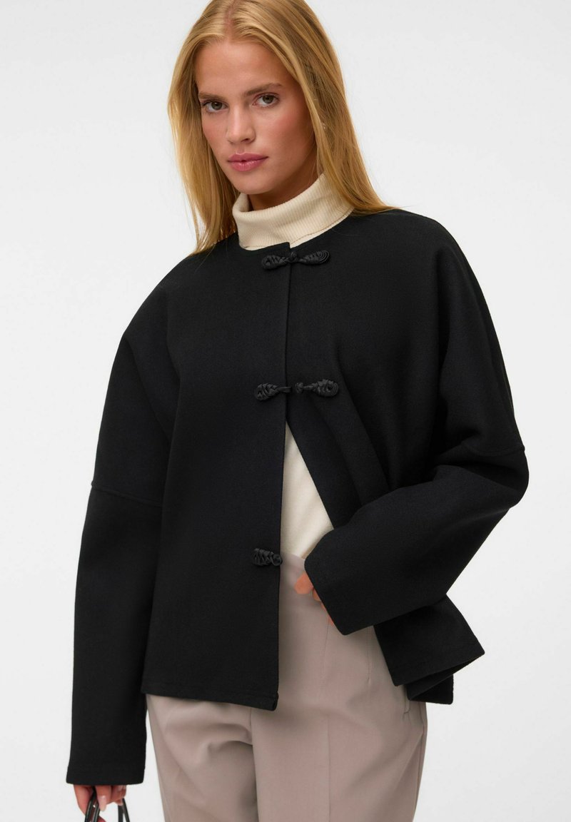 Femme blonde portant une veste noire avec trois fermetures à bascule sur un col roulé blanc et un pantalon beige, tenant un petit objet noir.