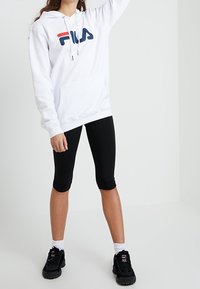 Felpa bianca con logo FILA rosso e blu, tasca frontale, abbinata a leggings neri e sneakers neri, indossata con calzini bianchi.