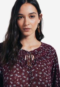 Blusa floreale bordeaux con scollo arricciato e dettaglio alla cintura, caratterizzata da motivi blu chiaro e bianchi. Tessuto morbido e trasparente con un design fluttuante.