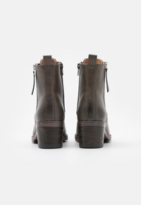 Un par de botas de tobillo de cuero marrón oscuro con tacones de bloque de madera y cremalleras laterales, vistas desde atrás sobre un fondo blanco.