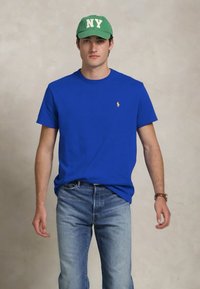 Jovem usando um boné verde de NY, t-shirt azul, calças jeans claras e um relógio, posando contra um fundo neutro.