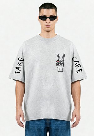 Homme portant un t-shirt gris oversize avec du texte noir "TAKE" sur la manche gauche, "CARE" sur la manche droite, et un graphisme de signe de paix de la main sur la poitrine.