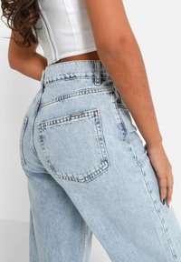 Donna che indossa jeans a vita alta azzurri chiari e un top corto bianco, mostrata di schiena e di lato su uno sfondo uniforme.