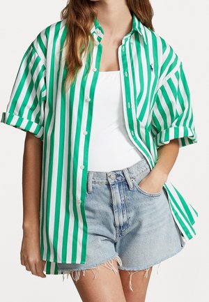 Vrouw draagt een oversized groen-wit gestreept overhemd met verticale knopen, een wit tanktop en lichtblauwe denim shorts met rafelige randen.