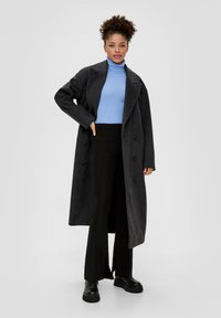 Langer grauer Wollmantel mit großen Revers, doppelt geknöpft, getragen über einem hellblauen Rollkragenpullover, kombiniert mit schwarzen Schlaghosen und klobigen Stiefeln.