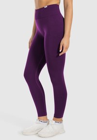 Lila hoch geschnittene Leggings aus dehnbarem Stoff, mit einer glatten Textur und ohne sichtbare Muster, kombiniert mit weißen Sportschuhen.