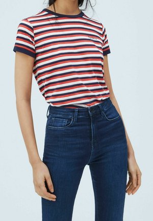 Vrouw die een donkerblauw, wit en rood gestreept T-shirt met korte mouwen draagt, ingestopt in een donkerblauwe spijkerbroek met hoge taille, tegen een effen achtergrond.