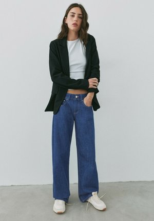Femme portant un blazer noir, un top court blanc, un jean large bleu et des baskets blanches, debout contre un mur clair et uni.