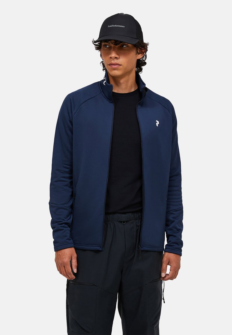 Marineblå zip-up jakke med høj krave, lavet af glat stof, med et hvidt logo på brystet og sorte elastiske manchetter.