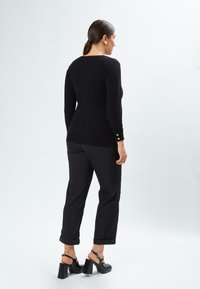 Schwarzer gestrickter Pullover mit langen Ärmeln und goldenen Knopfverzierungen an den Bündchen, kombiniert mit schwarzen, hochgerollten Hosen und klobigen Sandalen mit Absatz.
