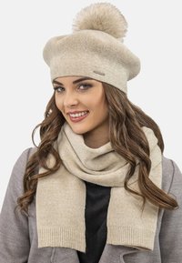 Bézs fonott beret, bolyhos pom-pommal és hozzáillő bézs sállal. A sál bordázott szélekkel és puha, textúrált felülettel rendelkezik.