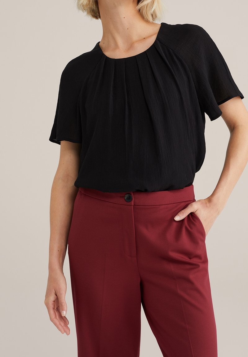 Femme portant un chemisier noir à manches courtes rentré dans un pantalon bordeaux taille haute, une main dans la poche, fond neutre.