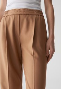 Pantaloni beige su misura con cintura elastica, caratterizzati da una texture liscia, una parte anteriore plissettata e tasche laterali.