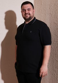 Čierna polo košeľa s trojvýpustkovým zapínaním, sivé akcenty na golieri a krátkymi rukávmi. Hladká textúra látky; logo sa nachádza na hrudi.