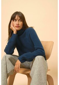 Modrý rolák z měkkého materiálu s žebrovanými manžetami, spárovaný se světle šedými pruhovanými kalhotami. Osoba sedí na dřevěné židli.