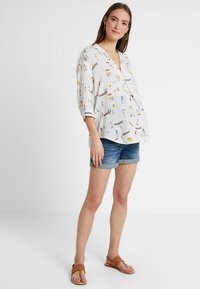 Blusa branca com padrões impressos coloridos, apresentando um decote em V e mangas três quartos. Combinada com calções de ganga azuis e sandálias castanhas.