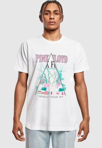 T-shirt en coton blanc avec un graphisme Pink Floyd en rose, turquoise et bleu, présentant des motifs triangulaires et le texte "TOUR MONDIAL '87".